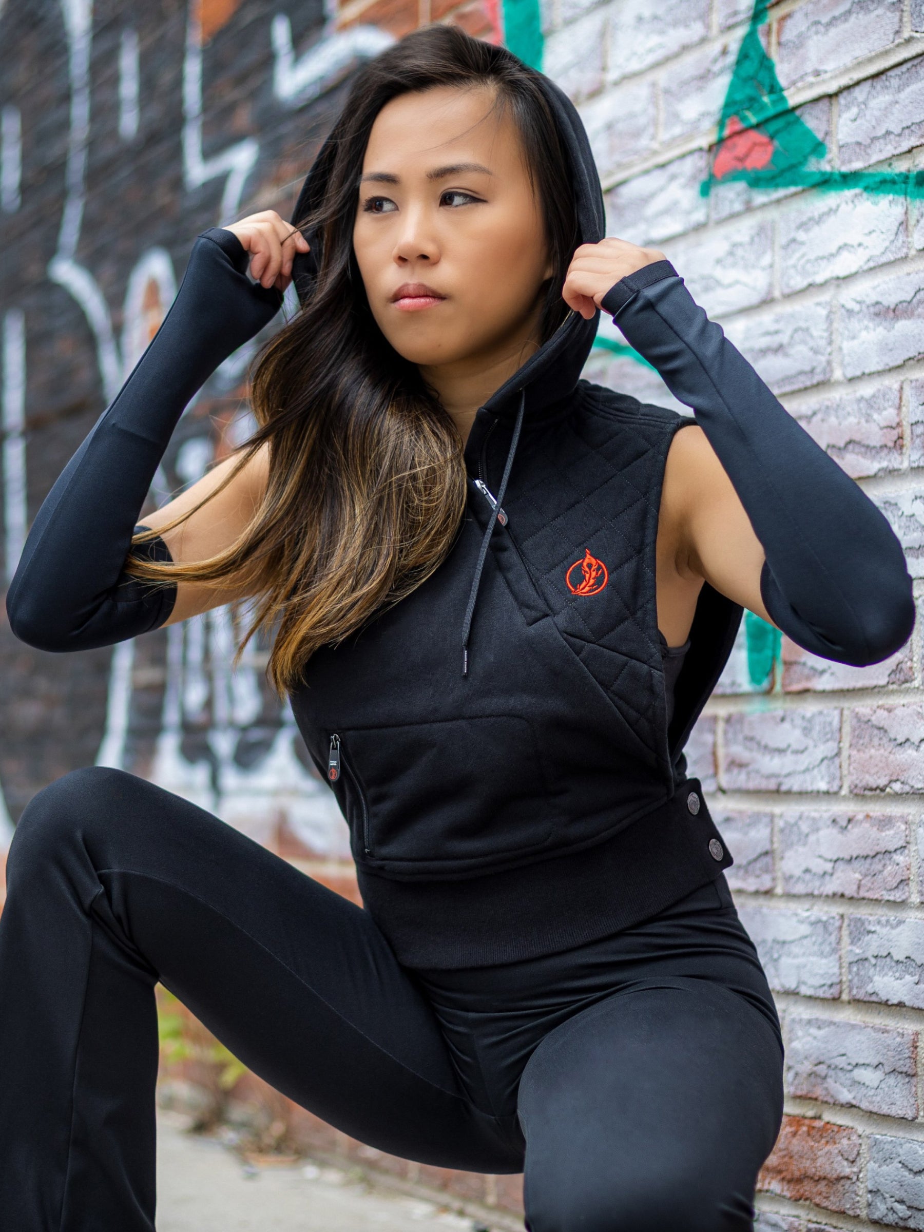 Model wearing Martialist Hoodie by Patch Notes Apparel at a 45-degree angle, highlighting diagonal zipper, padded quilting, main zip pocket, hem snaps, and sleeve gloves, with the model mid-way through adjusting the hood.